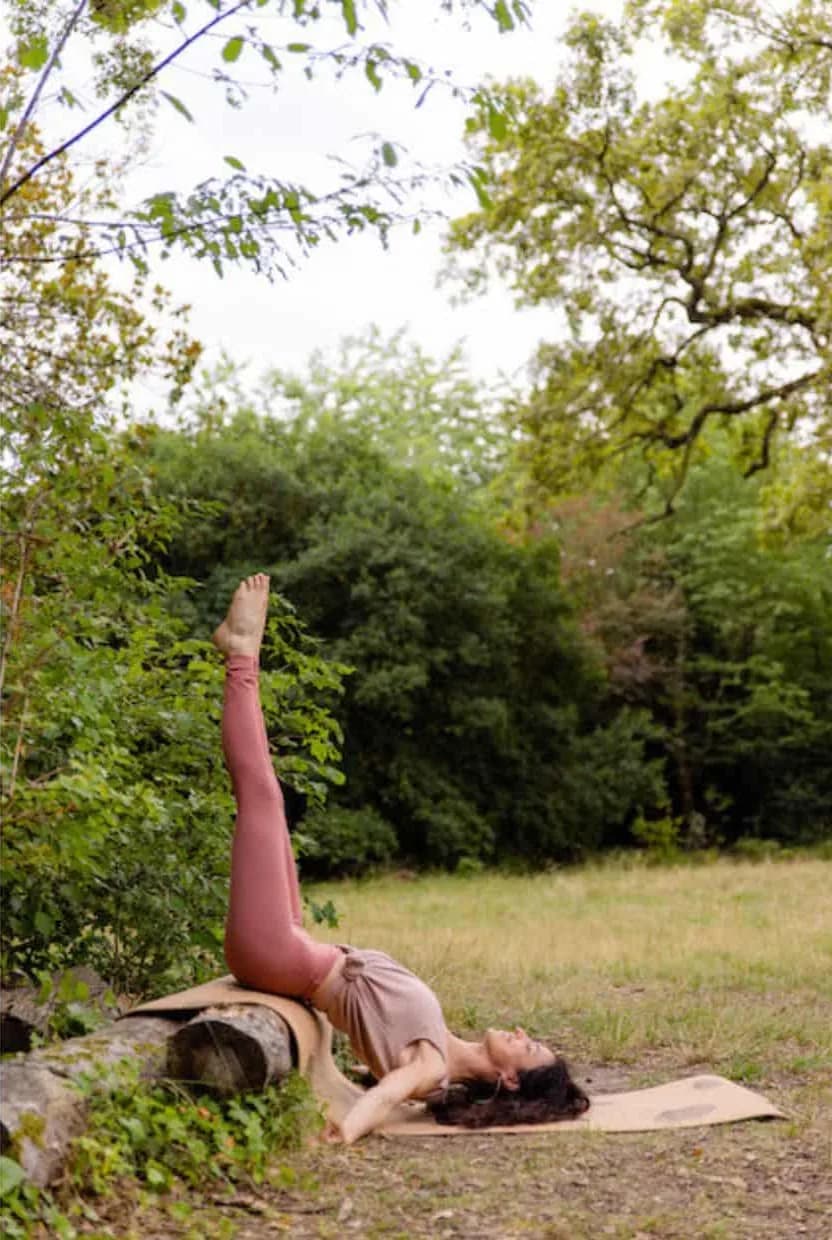 Une professeure de yoga allongée sur un tapis en liège dans une prairie, les jambes tendues vers le ciel